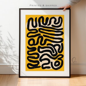 May include: Abstract art print in a black frame. The artwork features black, curved lines on a yellow background with off-white shapes. The text "PRINTED & SHIPPED" is at the top. The print is held by a person.