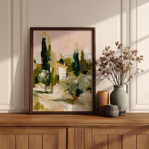 Puede incluir: Un cuadro de paisaje enmarcado que muestra altos cipreses de color verde oscuro, un peque&ntilde;o edificio blanco y follaje verde oliva suave sobre un cielo rosa p&aacute;lido. Junto al cuadro hay un jarr&oacute;n de cer&aacute;mica gris con flores blancas secas.