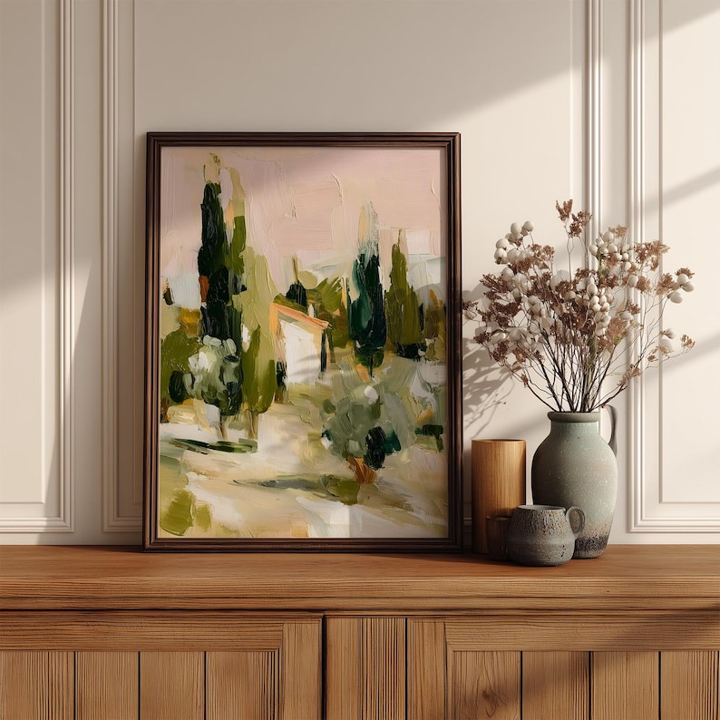 Puede incluir: Un cuadro de paisaje enmarcado que muestra altos cipreses de color verde oscuro, un peque&ntilde;o edificio blanco y follaje verde oliva suave sobre un cielo rosa p&aacute;lido. Junto al cuadro hay un jarr&oacute;n de cer&aacute;mica gris con flores blancas secas.