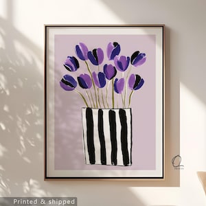 Può includere: Stampa artistica incorniciata con un bouquet di tulipani viola in un vaso a righe bianche e nere su sfondo lilla chiaro. L'opera d'arte ha uno stile moderno e minimalista, con colori audaci e forme semplici. La stampa è in una cornice dorata.