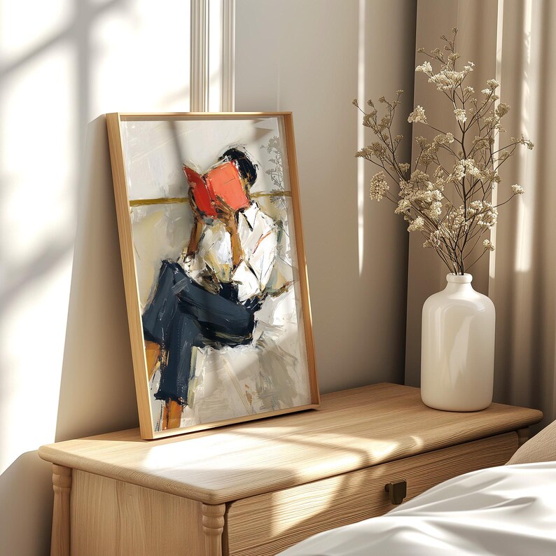 May include: A framed art print depicts a person absorbed in reading a book. The artwork features a person in a white shirt and dark pants, with a bright red book. The frame is light wood, and the painting has a modern, abstract style.