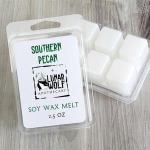 Op de afbeelding: Een doorzichtige plastic verpakking met zes witte sojawasblokjes. Het etiket bevat de tekst SOUTHERN PECAN, LUNAR WOLF APOTHECARY en 2.5 OZ, samen met een zwart-witte afbeelding van een rokende ketel en sterren.