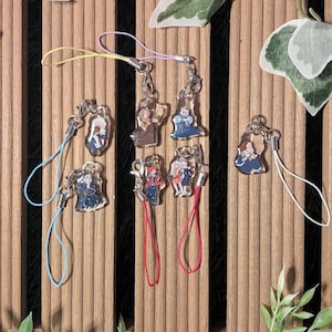 May include: A collection of acrylic charms featuring anime-style character designs. Each charm is attached to a metal clasp and a colorful lanyard. The charms are displayed on a wooden background with decorative greenery.