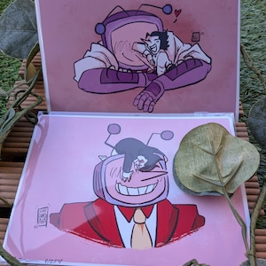 May include: Two art prints featuring cartoon characters. One print shows a purple robot embracing a smaller character, with a heart above them. The other print depicts a smiling robot in a red suit. The prints are on pink backgrounds.