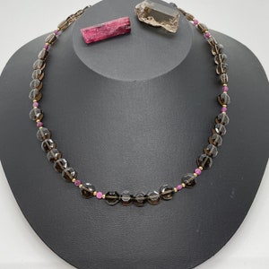 May include: A brown gemstone necklace with gold accents and small pink beads. The necklace is displayed on a black velvet stand.