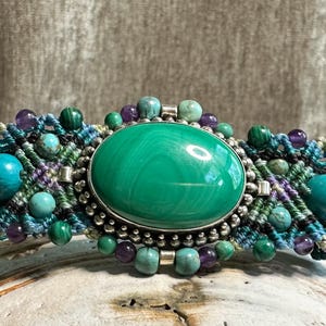 May include: A handcrafted bracelet featuring a large, oval malachite stone at the center, surrounded by smaller turquoise, amethyst, and malachite beads. The macrame band is in shades of blue, green, and purple.