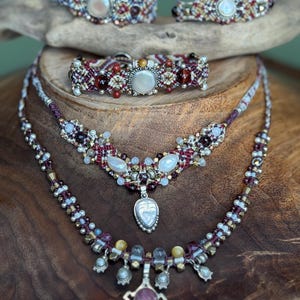May include: A collection of handcrafted macrame jewelry featuring two necklaces and three bracelets. The pieces are woven with deep red and neutral threads, accented with various gemstones, metallic beads, and central cabochon stones in silver settings.