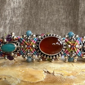 May include: A handcrafted bracelet featuring a central oval carnelian stone, flanked by turquoise and amethyst accents. The bracelet is intricately woven with colorful threads and adorned with silver-toned beads, creating a bohemian style.