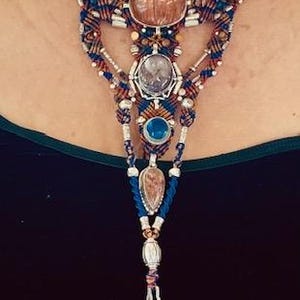 May include: A multi-strand necklace with a large, faceted, pink stone at the center. The necklace is made of brown, blue, and silver beads and cord. The necklace has a long, beaded tassel.