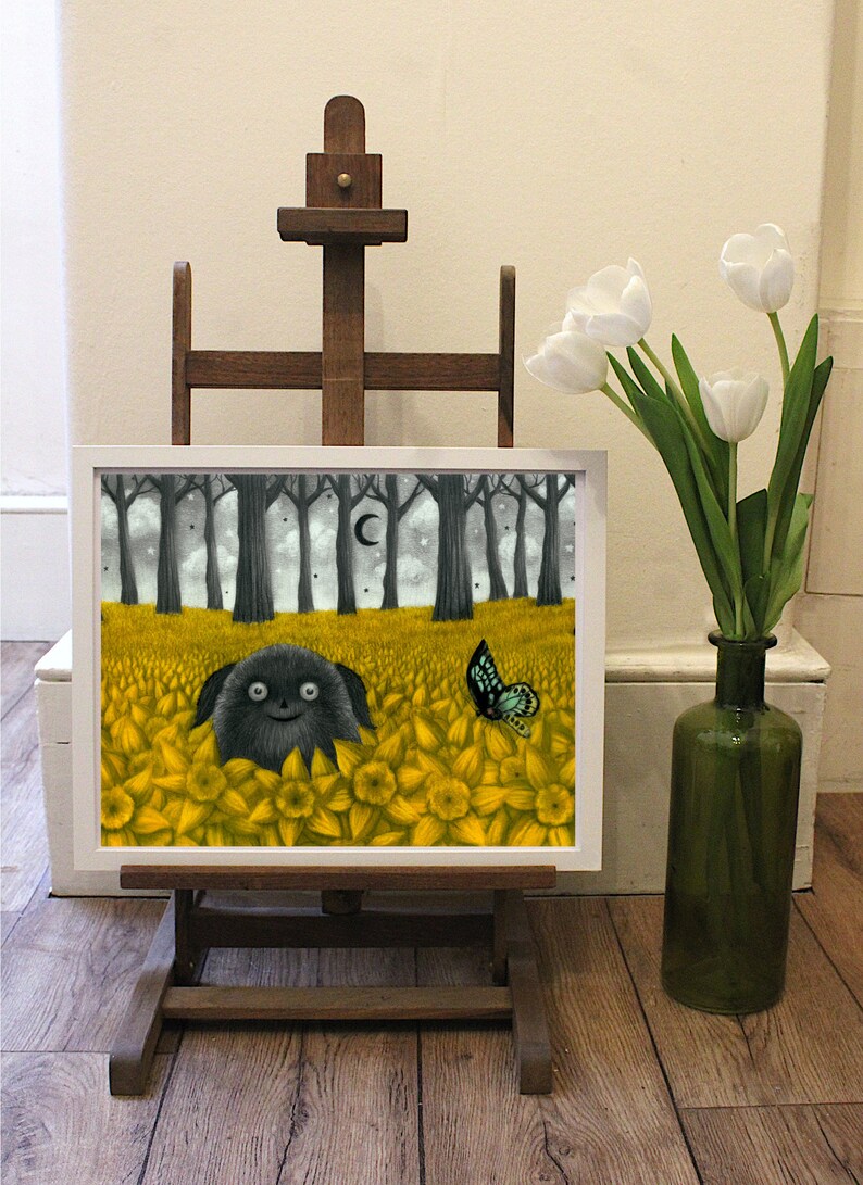 May include: A framed print of a black and white illustration of a creature with large eyes and a wide smile, surrounded by yellow flowers. A blue butterfly is flying above the creature. The print is on an easel in front of a white wall.