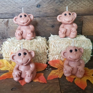 May include: Four brown monkey candles sitting on hay bales. The monkeys are sitting with their legs crossed and have a happy expression on their faces.