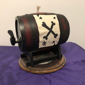 Op de afbeelding: Een decoratief vat met een zwarte en rode afwerking, met een kraan en een witte banner met een zwart doodshoofd en gekruiste botten ontwerp. Het vat staat op een houten voet en standaard, wat een piratenthema oproept.