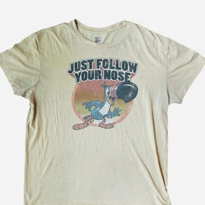 Könnte beinhalten: Beigefarbenes T-Shirt mit einem Cartoon-Tukan und dem Text "JUST FOLLOW YOUR NOSE". Der Tukan ist blau und rot, mit einem großen schwarzen Schnabel, vor einem roten und orangefarbenen Kreis. Das T-Shirt hat einen Vintage-Look.