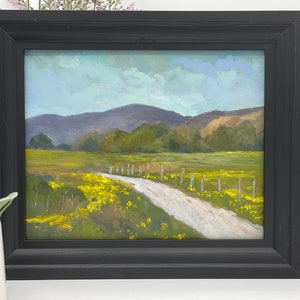 May include: A framed oil painting depicting a rural landscape with a dirt road leading through a field of yellow wildflowers. The road is bordered by a wooden fence and the background features rolling hills and a blue sky with white clouds.