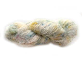 Puede incluir: Una madeja de hilo esponjoso de color crema con una textura suave y cepillada. El hilo está salpicado con sutiles y delicados puntos de fibras en azul pastel, amarillo pálido y rosa claro distribuidos por todo el haz de fibras retorcido.