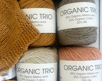 May include: Four skeins of yarn in different colours: mustard yellow, light grey, light pink, and grey. Each skein has a white label with the text "ORGANIC TRIO" and the yarn composition: 50% Organic Merino wool, 25% Organic Cotton, 25% Silk.