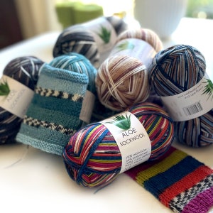 May include: A close-up of colorful yarn balls, including a skein of multi-colored yarn labeled "Aloe Sockwool" with the text "75% wool s/w 25% polyamid 100g-ca.420m 30m-10ТУРА 1277 www.njenegon.com sitting with love".