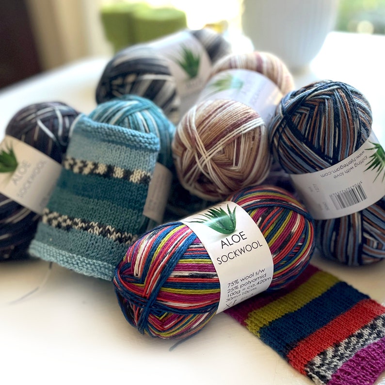 May include: A close-up of colorful yarn balls, including a skein of multi-colored yarn labeled "Aloe Sockwool" with the text "75% wool s/w 25% polyamid 100g-ca.420m 30m-10ТУРА 1277 www.njenegon.com sitting with love".