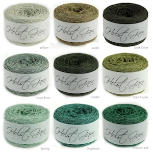 Puede incluir: Nueve ovillos de lana cil&iacute;ndricos dispuestos en cuadr&iacute;cula, cada uno envuelto en una banda de papel blanco con la etiqueta Holst Garn. Los colores incluyen tonos verde claro, oliva, salvia, verde oscuro y azul verdoso, con los nombres Willow, Heath, Dark Olive, Sage Blue, Tundra, Dark Apple, Spring, Kingfisher y Clover Leaf impresos debajo de cada uno.