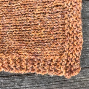 Puede incluir: Una vista de cerca de una tela tejida a mano con un patr&oacute;n de punto jersey en hilo de color marr&oacute;n &oacute;xido jaspeado. El tejido muestra una superficie texturizada con bucles de fibra visibles y un tejido denso y consistente en toda la pieza.