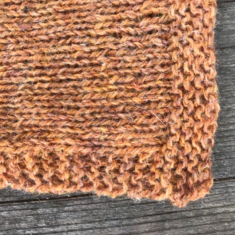 Puede incluir: Una vista de cerca de una tela tejida a mano con un patr&oacute;n de punto jersey en hilo de color marr&oacute;n &oacute;xido jaspeado. El tejido muestra una superficie texturizada con bucles de fibra visibles y un tejido denso y consistente en toda la pieza.