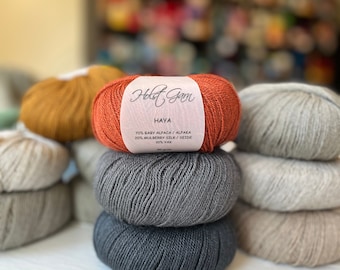 Puede incluir: Una pila de ovillos de lana en varios colores, incluyendo naranja, gris y beige. El ovillo superior tiene una etiqueta que dice "Hold Gaon HAYA 70% Baby Alpaca/Alpaka 20% Mulberry Silk/Seide 10% Yak".