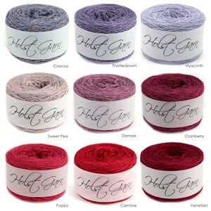 Puede incluir: Nueve ovillos de lana cil&iacute;ndricos dispuestos en cuadr&iacute;cula, cada uno envuelto en una banda de papel blanco con la etiqueta Holst Garn. Los colores incluyen tonos de azul, morado, lavanda, beige, rosa, magenta y rojo oscuro, etiquetados como Crocus, Thistledown, Hyacinth, Sweet Pea, Damask, Cranberry, Poppy, Carmine y Venetian.