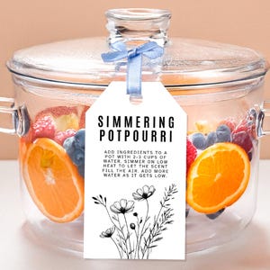 May include: A clear glass pot filled with fruit slices and berries, with a tag that reads "Simmering Potpourri." The tag includes instructions and a floral illustration. A blue ribbon secures the lid.