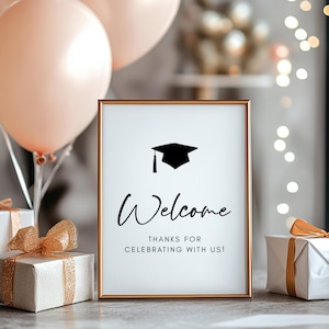 Könnte beinhalten: Ein gerahmtes Schild mit goldenem Rand mit der Aufschrift „Willkommen“ und einer Abschlusskappen-Ikone. Das Schild ist von Geschenken umgeben, die in weißes Papier mit goldenen Schleifen und pfirsichfarbenen Luftballons verpackt sind. Ein Thema für eine Abschlussfeier.