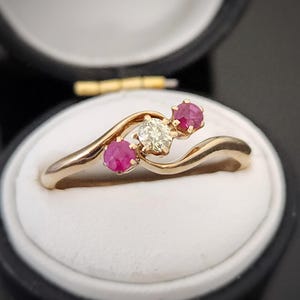 May include: A gold ring featuring a diamond and two ruby stones. The ring is displayed in a black ring box.