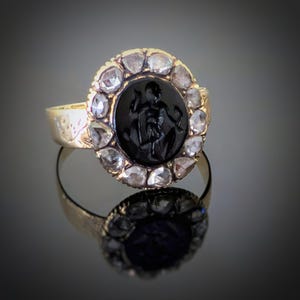 Könnte beinhalten: Ein goldener Ring mit einem schwarzen, ovalen Stein mit einer eingravierten Figur, umgeben von klaren, unregelmäßig geformten Steinen. Der goldene Ring und die Fassung fangen das Licht ein und erzeugen einen schimmernden Effekt. Der Ring wird auf einer reflektierenden Oberfläche präsentiert.