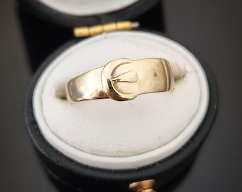 May include: A gold ring with a buckle design. The ring is in a black ring box.