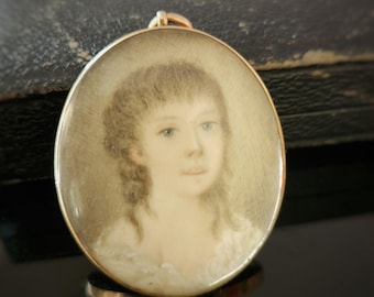 May include: Oval gold pendant featuring a miniature portrait of a person with light skin, blue eyes, and light brown hair. The portrait is set within a gold frame, showcasing a delicate painting style. The pendant is likely vintage or antique.