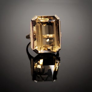 May include: A gold ring featuring a large, rectangular, emerald-cut, translucent brown gemstone set in a four-prong mounting. The stone displays internal facets and light reflections, with the entire piece casting a clear reflection below it.