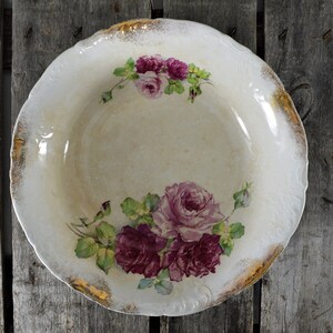 May include: A white ceramic bowl with a gold rim and floral design. The bowl features pink and purple roses with green leaves.