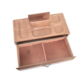 Peut inclure: Un organiseur de bureau en bois avec un tiroir coulissant et plusieurs compartiments. La partie supérieure comporte diverses zones encastrées pour ranger de petits objets. Le tiroir est doublé d'un tissu brun-rosé. L'organisateur est en bois clair.