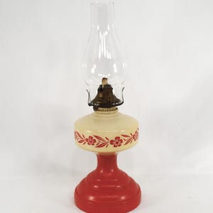 May include: A vintage oil lamp with a clear glass chimney and a cream-colored body. The body is decorated with red floral patterns and sits atop a red base. The lamp is designed for illumination.