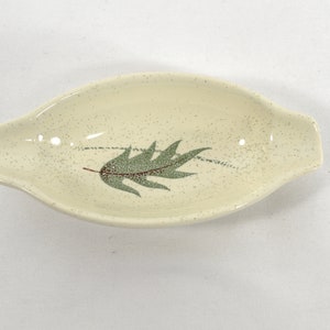 May include: A small, white ceramic dish with a green leaf design. The dish has a speckled glaze and a slightly oval shape.