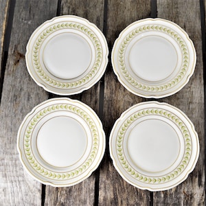 May include: Four white ceramic plates with a gold rim and green leaf design. The plates are arranged in a square pattern on a wooden surface.