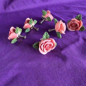 May include: A set of six drawer knobs shaped like pink roses with green leaves. Each ceramic rose features a metal screw attachment extending from the back, designed for installation on furniture or cabinetry hardware.