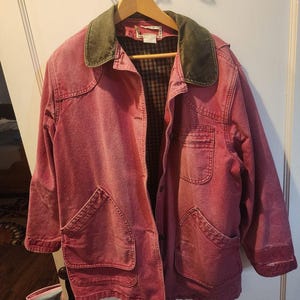 May include: A faded red denim jacket featuring a dark olive green corduroy collar and a button-front closure. The jacket includes two large front patch pockets, a single chest pocket, and a dark plaid interior lining, all hanging on a wooden hanger.
