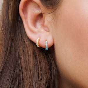 May include: A woman wearing a pair of gold hoop earrings with small gemstones. One earring has white gemstones and the other has blue gemstones. She is also wearing a gold ring on her finger.