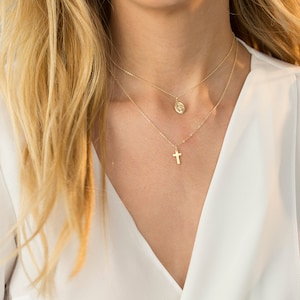 May include: Two gold necklaces with a cross pendant and a round pendant. The necklaces are layered on a person's neck.