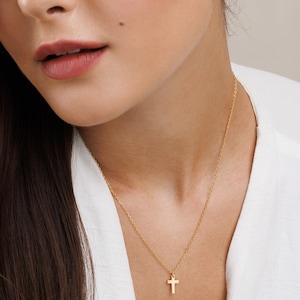 May include: A gold chain necklace with a small gold cross pendant. The necklace is worn by a person with long brown hair and a white shirt. The person is also wearing a pair of pearl earrings.