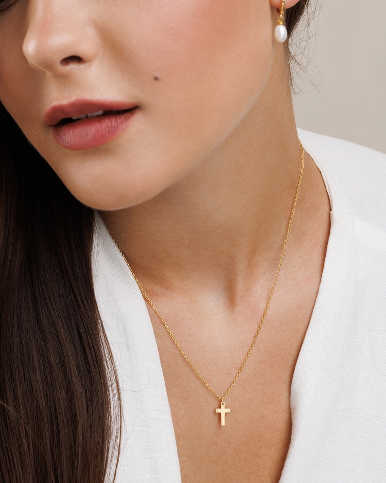 May include: A gold chain necklace with a small gold cross pendant. The necklace is worn by a person with long brown hair and a white shirt. The person is also wearing a pair of pearl earrings.