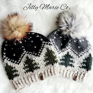 Op de afbeelding: Twee gebreide wintermutsen met nep bont pompoenen. De ene muts is zwart, wit en groen met een bruine nep bont pompon. De andere muts is grijs, wit en groen met een witte nep bont pompon. Beide mutsen hebben een bruin lederen label met de tekst "Jilly Marie Co."
