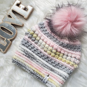May include: A gray, pink, and yellow striped crocheted beanie with a fluffy pink pom pom. The beanie has a small tag that says "Be Kind".