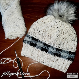 May include: A crocheted beanie hat with a white and black checkered band and a fluffy gray pom pom. The hat is made with a chunky yarn and has a textured stitch pattern. A ball of white yarn and a crochet hook are also in the image.