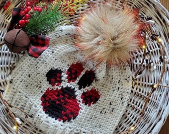 May include: White crocheted beanie with a red and black buffalo plaid paw print design. The beanie has a fluffy brown pom pom and a brown leather tag with the initials "JMC".
