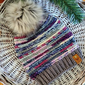 Könnte beinhalten: Eine bunte gestreifte Strickmütze mit einem Kunstfellbommel oben. Die Mütze hat einen braunen Umschlag und ein kleines braunes Leder-Etikett mit dem Wort "kind" darauf.